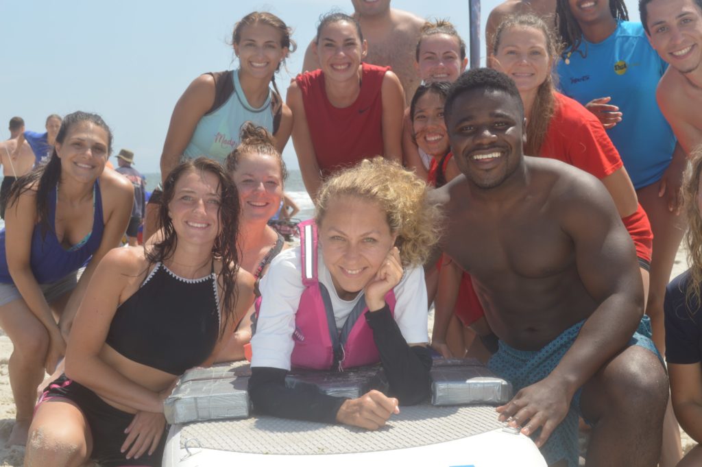 group of smiling young people surrounding a young woman laying on a surfboard.