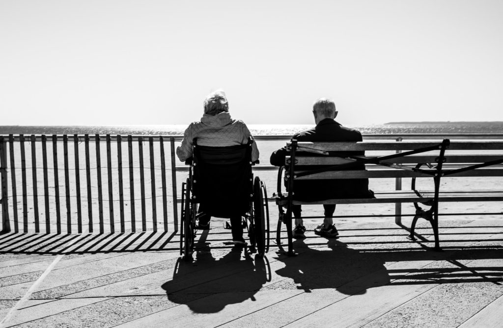 black and white image of an elderly couple sitting on a boardwalk behind a metal railing looking out over a beach. The woman is sitting in a wheelchair, the man is sitting next to her on a park bench. They are photographed from behind on a sunny day.