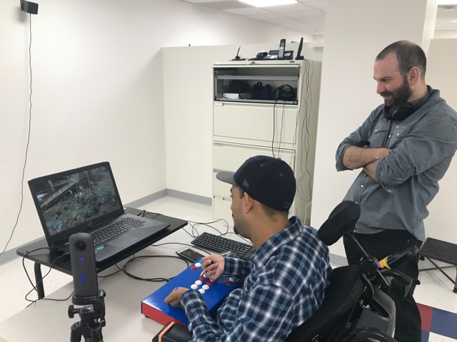 Young man in a wheelchair plays adaptive video game while young man standing behind him watches.