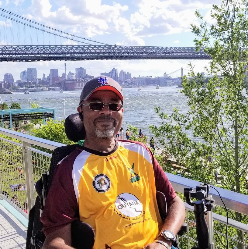 Image ID: Black man with a short gray goatee and moustache is smiling at the camera. He is sitting in a wheelchair outside on a sunny day, wearing a blue baseball cap, dark glasses and a red and yellow short sleave shirt. The RFK Bridge in the background.